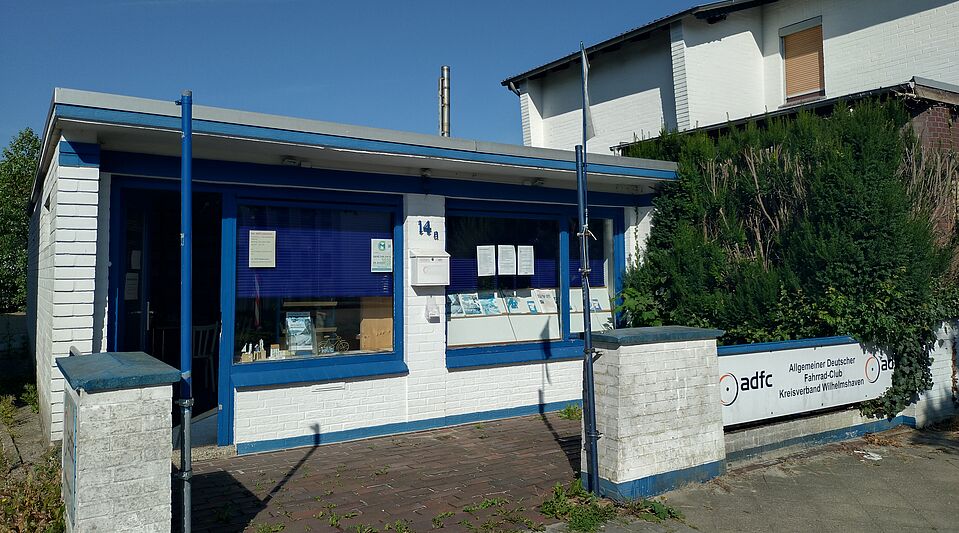 Infoladen Ein weißer, gemauerter Bungalow mit blauen Tür- und Fensterrahmen.