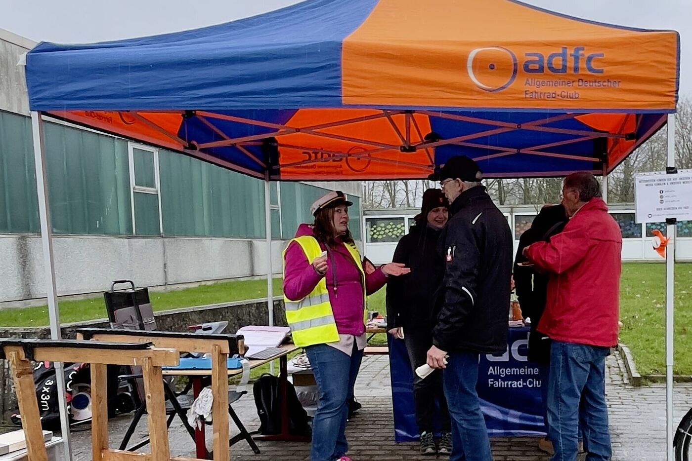 Vier Menschen stehen unter einem Pavillon an einem Infostand. Sie tragen Jacken. Sie unterhalten sich.