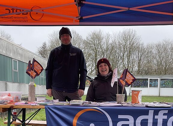 Zwei Menschen stehen unter einem Pavillon an einem Info-Stand. Sie schauen freundlich in die Kamera.