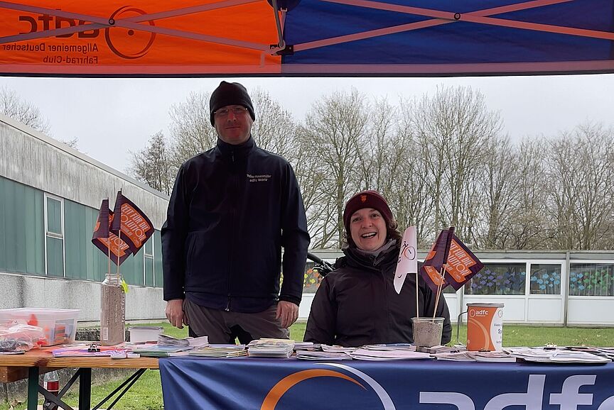 Zwei Menschen stehen unter einem Pavillon an einem Info-Stand. Sie schauen freundlich in die Kamera.